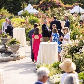 People mingling at high-top tables in a blooming rose garden.