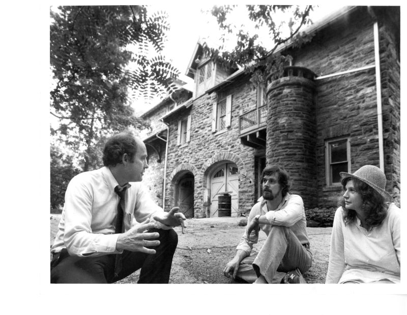 A black and white photo of a three people sitting on the ground talking outside of a stone building.