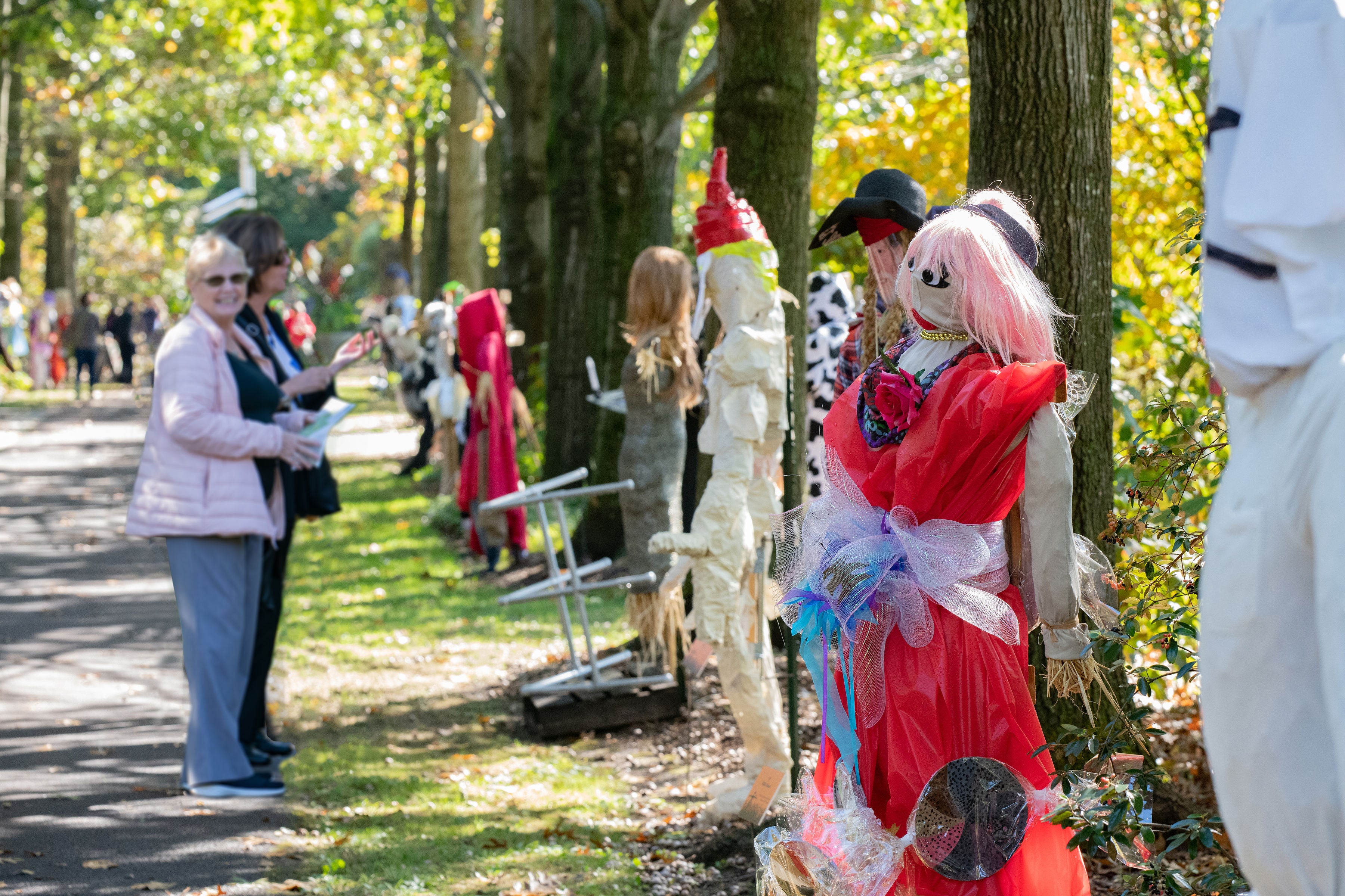 Two woman smile and look at a row of designed scarecrows in an outdoor setting.