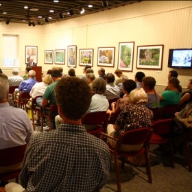 A group of people sit in chair indoors and listen to a speaker at a podium. 