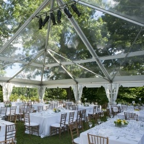 A clear-top wedding reception tent outside and surrounded by green trees.