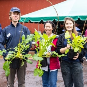 Three people stand in front of a red barn smiling and holding plants. 