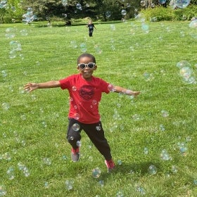 A young boy wearing sunglasses extends his arms to the sides while running through bubbles in a sunny green field.