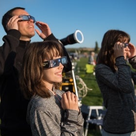 Two parents and a child look up at the sky wearing protective glasses to see a solar eclipse.