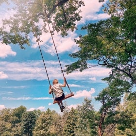 The back of a young child with blonde hair on a swing with a backdrops of blue sky, clouds, and trees. 