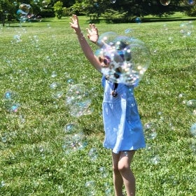 A young girl wearing sunglasses and jumping with her arms up through a field of bubbles. 