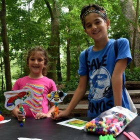 Two children smile for the camera while holding up insect crafts in an outdoor space. 