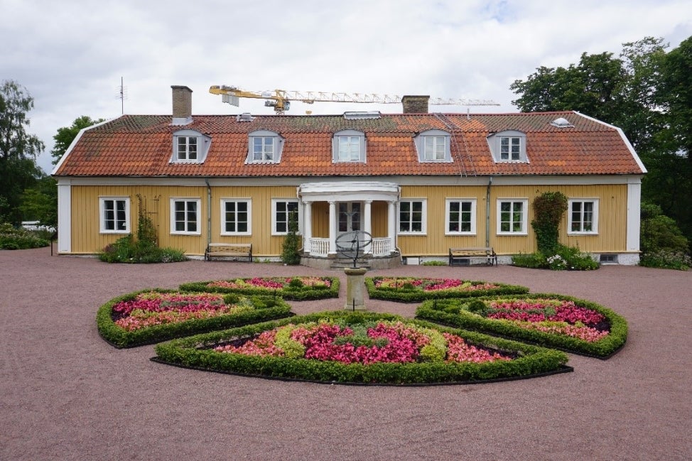 A yellow building in the background. In the foreground, a circle garden bed design dissected into five beds with pink flowers and green foliage. 