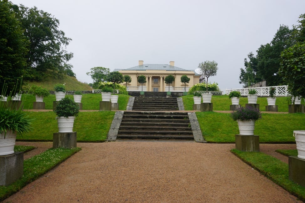 A large pillared building on top of a hill with concrete steps leading to the front and potted plants surrounding it. 