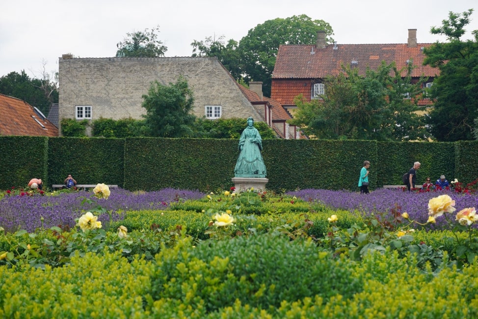 A large land garden filled with purple, red, and yellow flowers and green foliage, with a green statue of a woman in a dress in the middle and people walking around. 