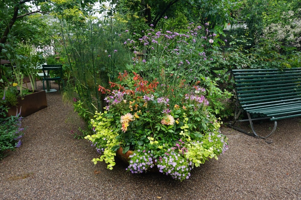 A large outdoor garden planter filled with colorful flowers and next to a green bench. 