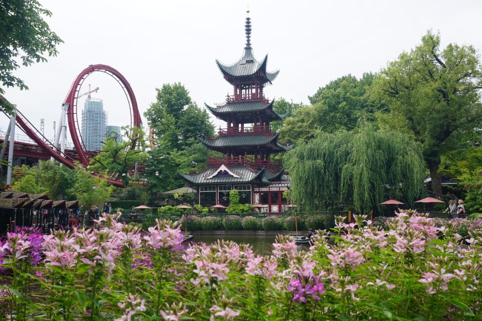 In the background is a large pagoda, rollercoaster, and body of water; in the foreground is pink and purple flowers. 