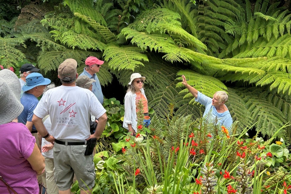 A group of people stand outdoors surrounded by flowers and large foliage while a woman gives them a tour.