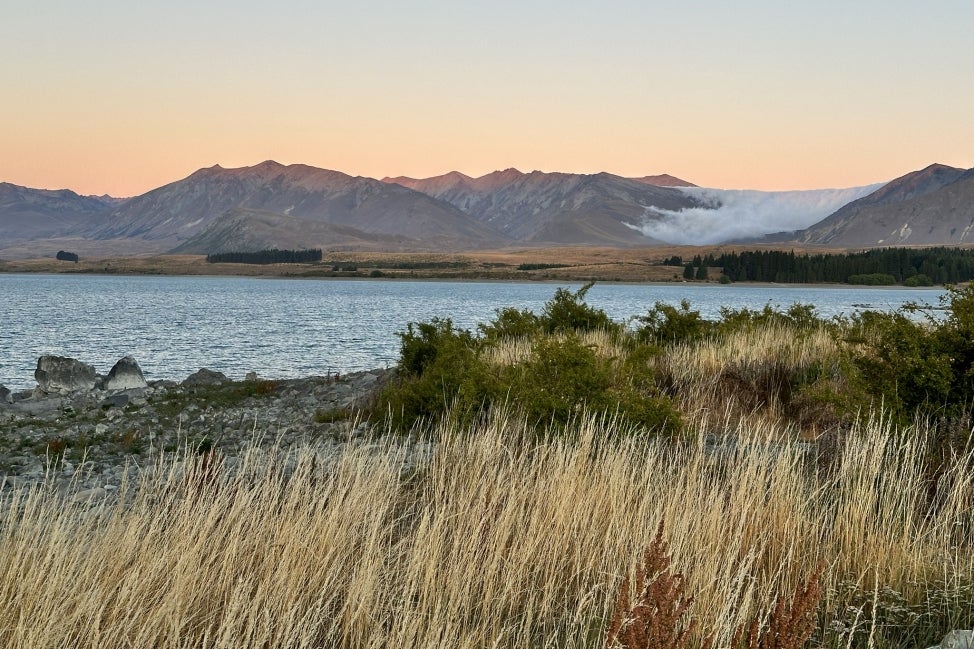 A grass landscape with a body of water and mountain in the background as the sun is setting. 