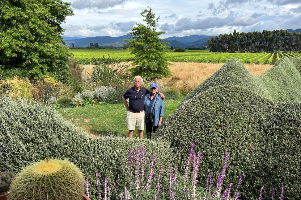 An older couple stands with their arms around each other outside in the middle of a green landscape filled with farms, foliage, and blooming flowers with mountains in the distance. 