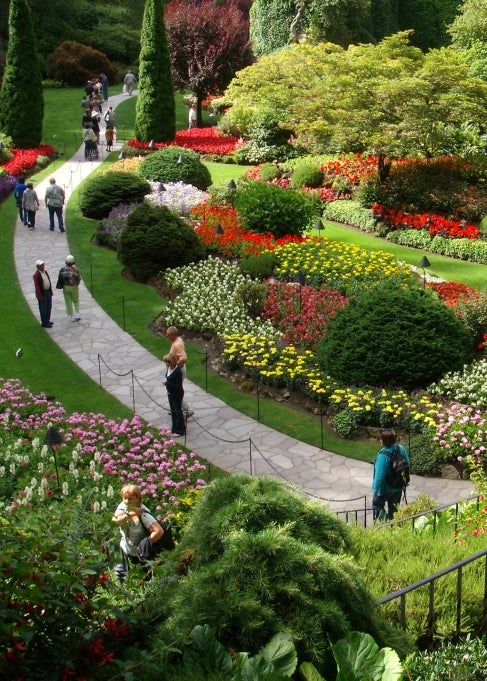 A public garden filled with blooming flower beds, trees, and other foliage. A stone path winds through it, filled with people. 