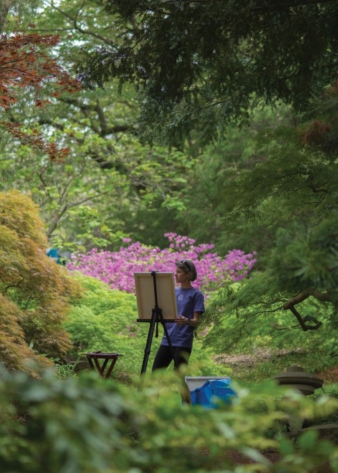 A woman stands painting at a easel outdoors in a garden with bright green foliage and a tree with pink flowers. 