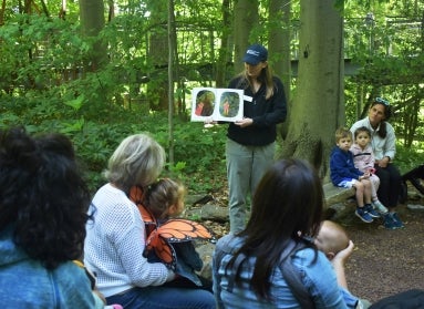 A young woman in a baseball cap reads a picture book to a group of children in a green, wooded setting. 