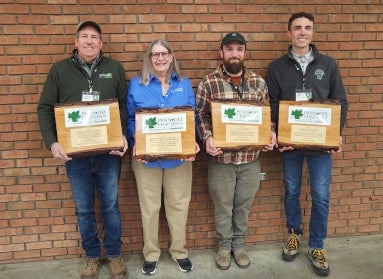 Four people stand in a row against a brick wall holding award plaques.