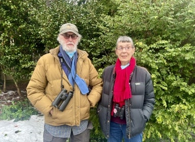 A man and a women wearing winter attire and binoculars stands in front of a green bush with snow on the ground.