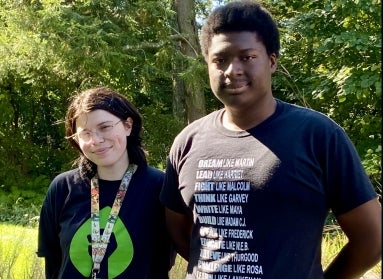 Two young students stand next to each smiling at the camera in a green outdoor setting. 