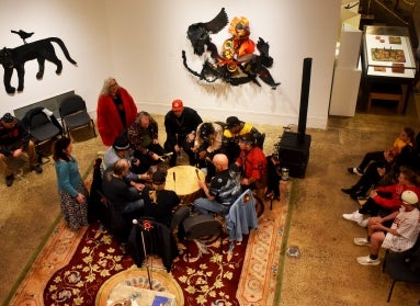 A group from the Lenape Nation of PA sit in a drum circle and perform in the middle of an art museum with an audience. 