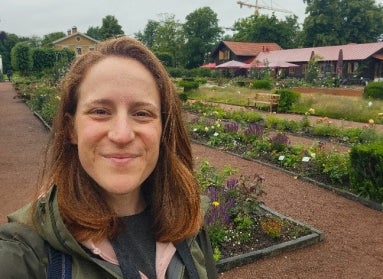 A young woman with red hair takes a selfie in front of long rows of garden beds. 