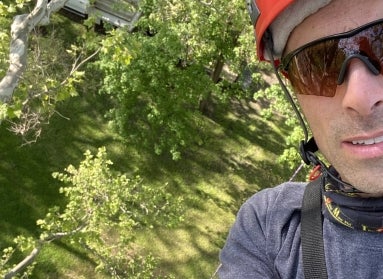 A man wearing sunglasses and a helmet high in a tree with a view of the ground below him