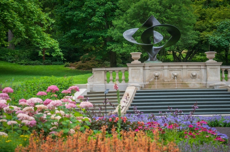 A looping metal sculpture at the top of a step fountain surrounded by pink, orange, and purple flowers. 