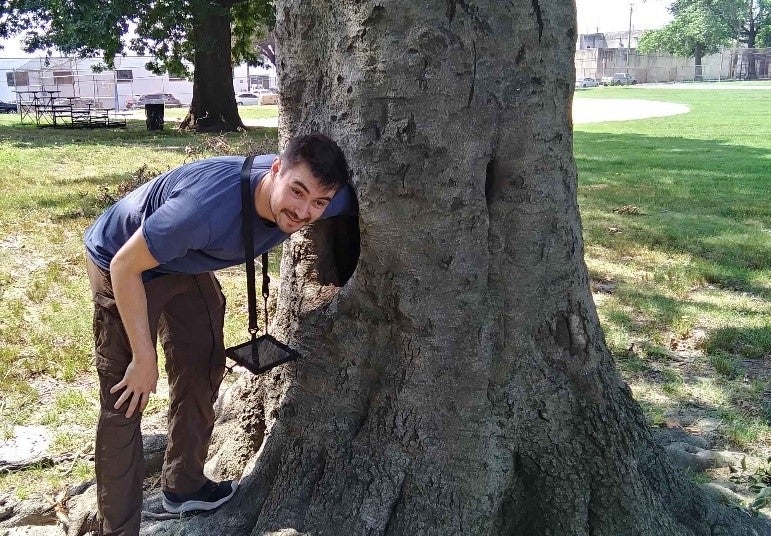 A young man stands with the trunk of a large tree. 