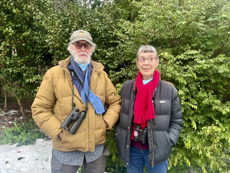 A man and a woman wearing winter attire and binoculars stand in front of green bush with snow on the ground.