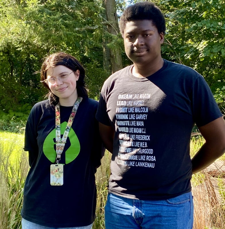 Two students stand next to each other smiling for the camera in a green outdoor setting. 