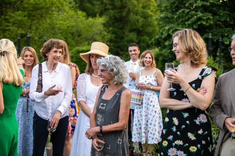 A group of people in garden party attire hold beverages and smile while talking outdoors. 