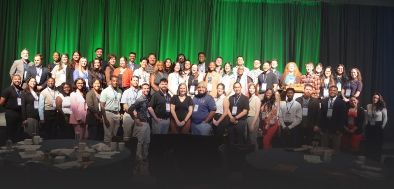 A large group of people standing on risers on a stage in front of an emerald green curtain.