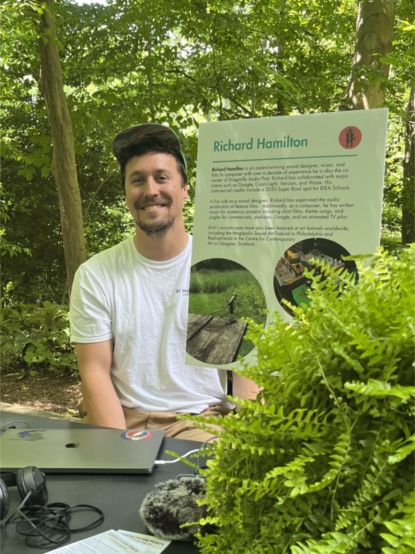 A man in a white t-shirt and baseball cap sits outdoors next to a sign that reads, "Richard Hamilton" followed by three paragraphs of small text. 