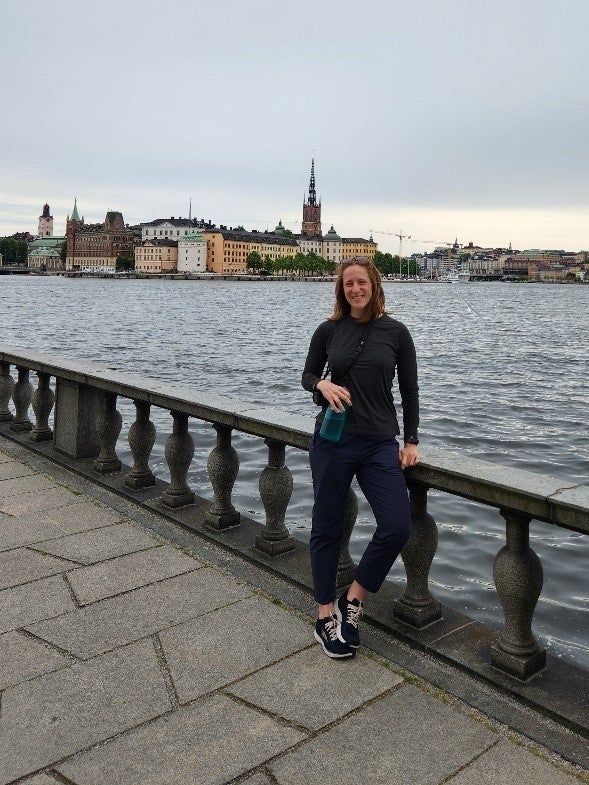 A woman leans against a cement railing overlooking a body of water and buildings in the distance. 