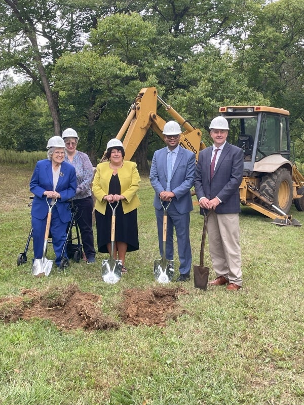 Five people in hardhats and holding shovels stand in front of a backhoe