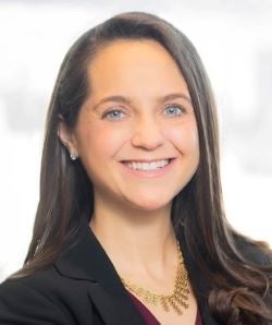 A headshot of a woman with long brown hair and blue eyes wearing a gold necklace and black blazer. 