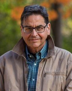 A portrait of a man with dark hair, eyeglasses, a blue checkered shirt, and a tan jacket. 