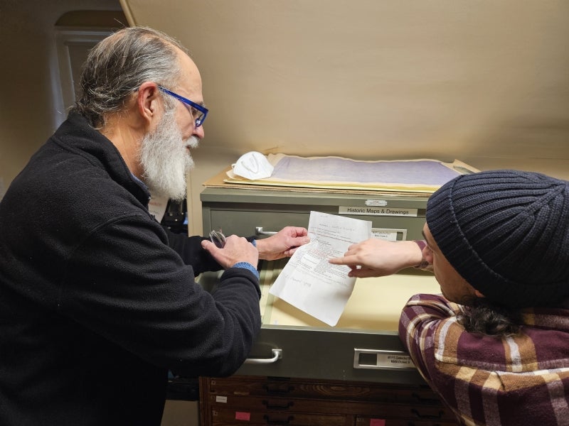 Two men stand in front of flat file. One holds a piece of paper while the other points at it. 