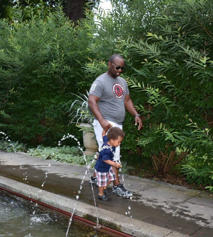 A man and his young son walk along a long fountain outdoors surrounded by green foliage. 